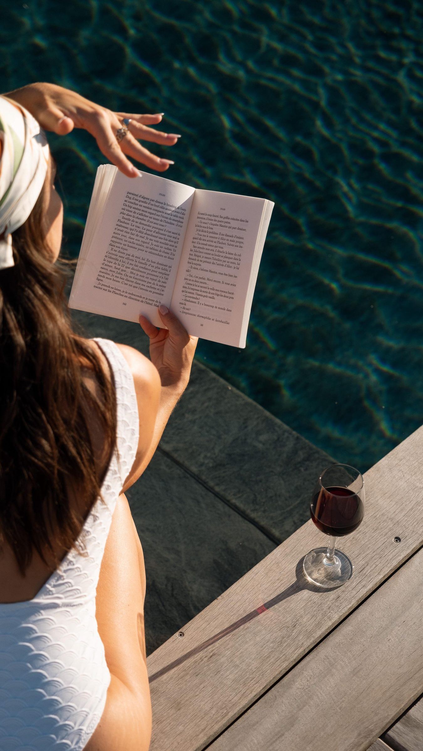 Une femme lit au bord de la piscine, un verre de vin rouge posé sur le bois